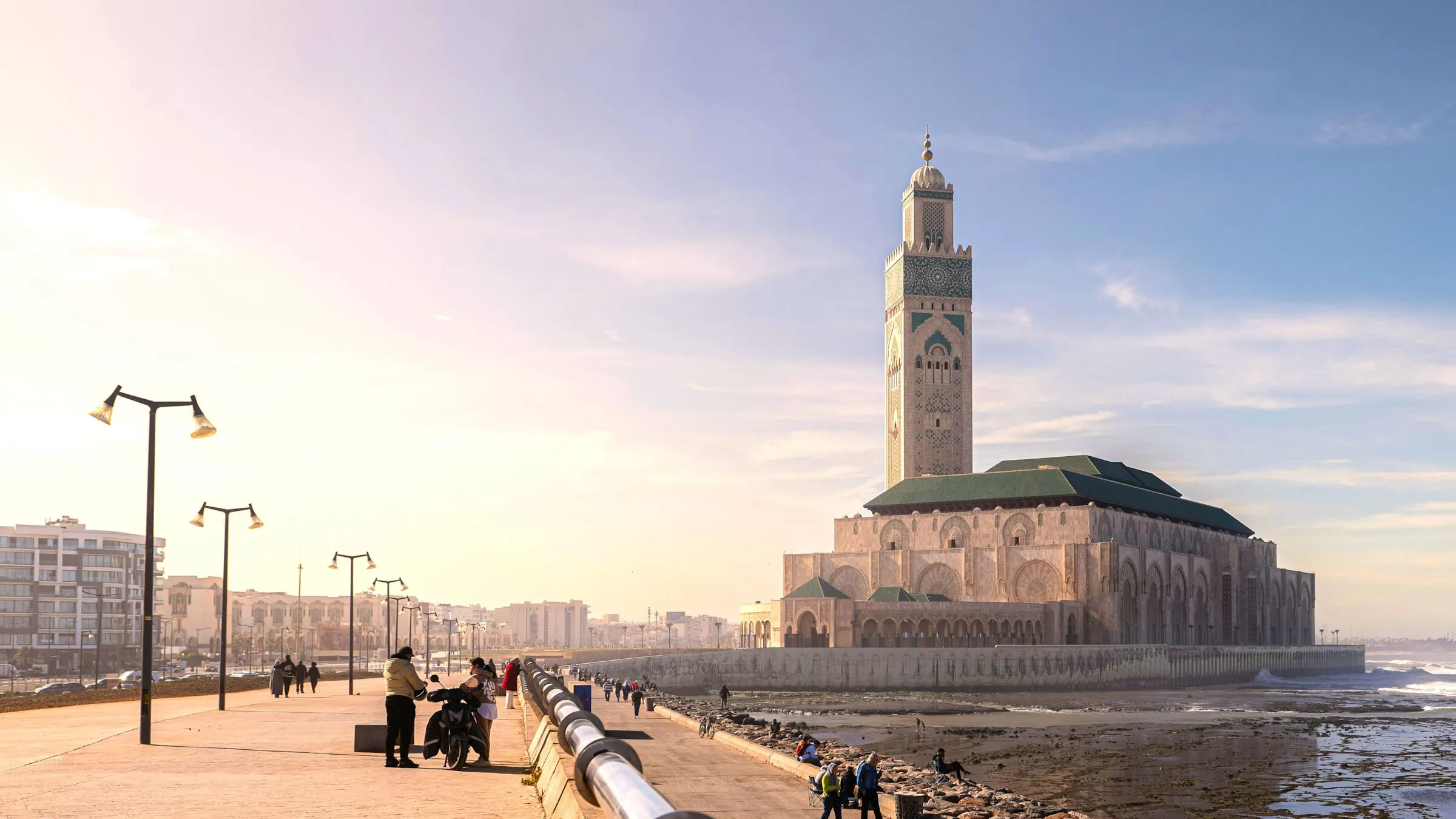 The Hassan II Mosque and its tall green-tiled minaret on the Casablanca seafront, seen from the corniche promenade at low tide